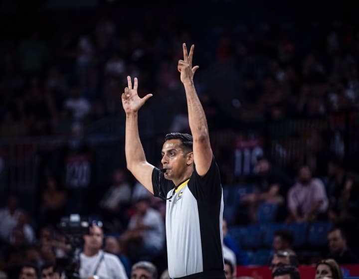 A referee signalling a three-point basket with both arms raised in a packed arena